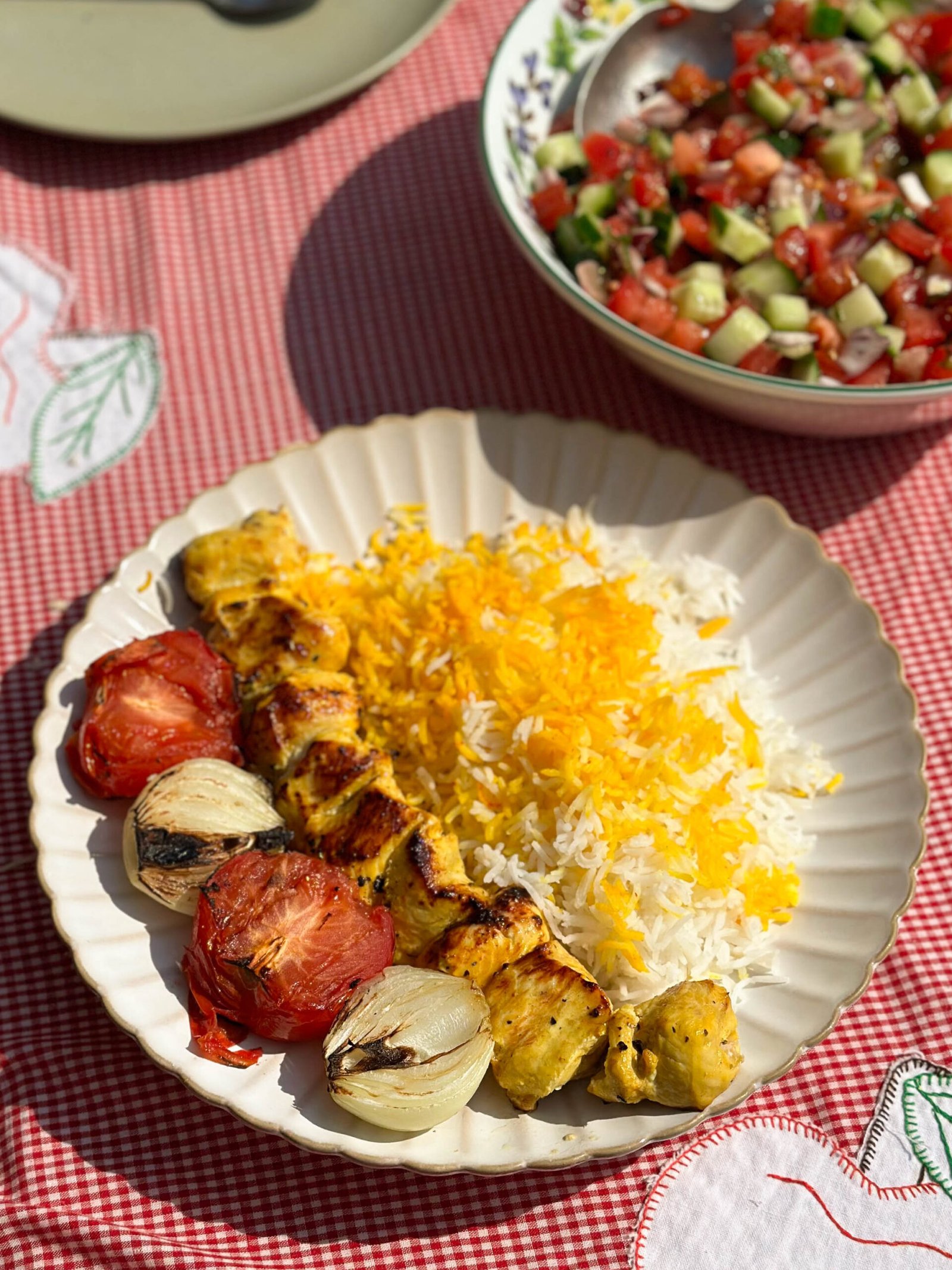 A delicious skewer of Joojeh KababPersian saffron chicken served with vibrant saffron rice, perfectly grilled tomatoes, and charred onions on a white plate.