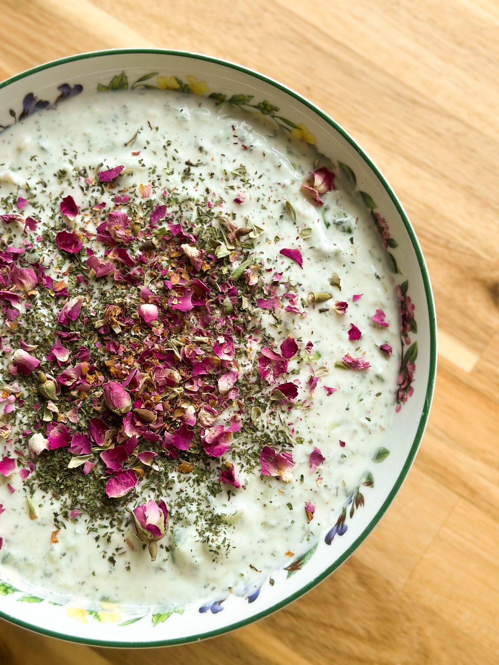 A bowl of creamy Mast-o-Khiar, persian yogurt salad, elegantly garnished with dried mint and delicate rose petals, on a decorative plate.