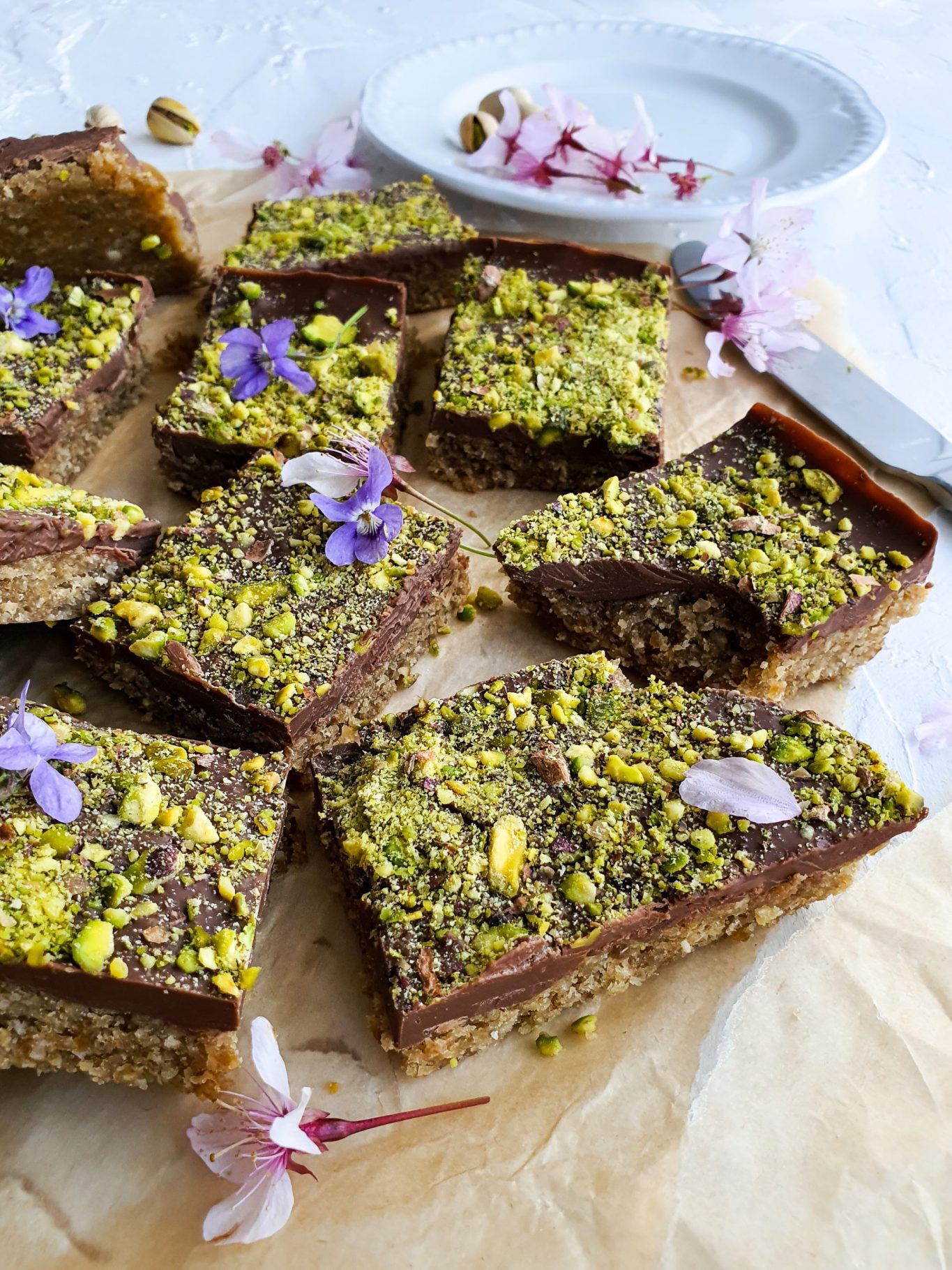 A stack of rectangular, golden-brown bars with a crumbly texture and visible chunks of green pistachios. The bars are sprinkled with a light dusting of powdered sugar.
