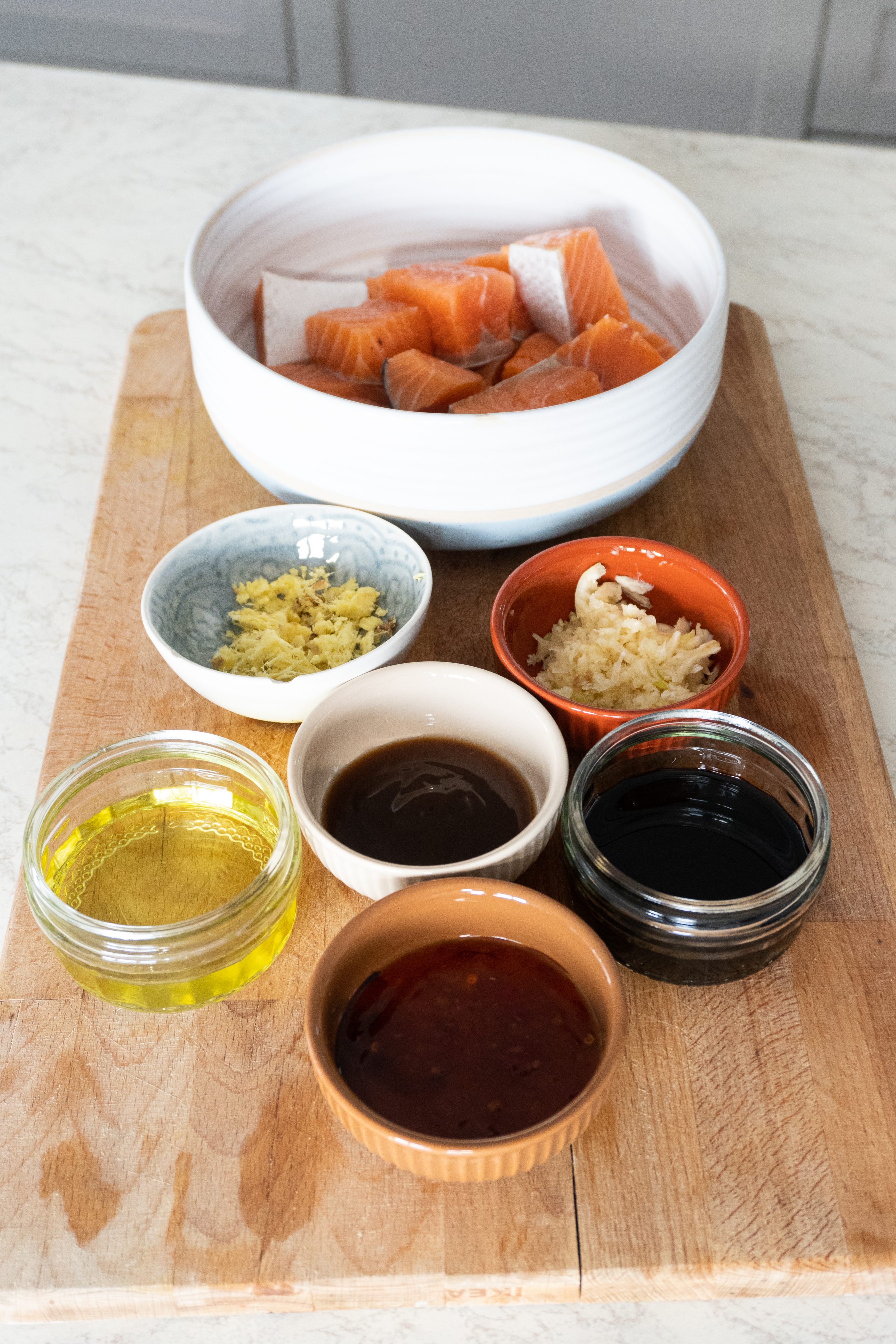 An image featuring the ingredients for a Crispy Honey Ginger Garlic Salmon Bowl. The photo shows a wooden cutting board with a selection of fresh ingredients, including salmon fillets, avocado, green onions, sesame oil, honey, soy sauce, oyster sauce, oil ginger and garlic.