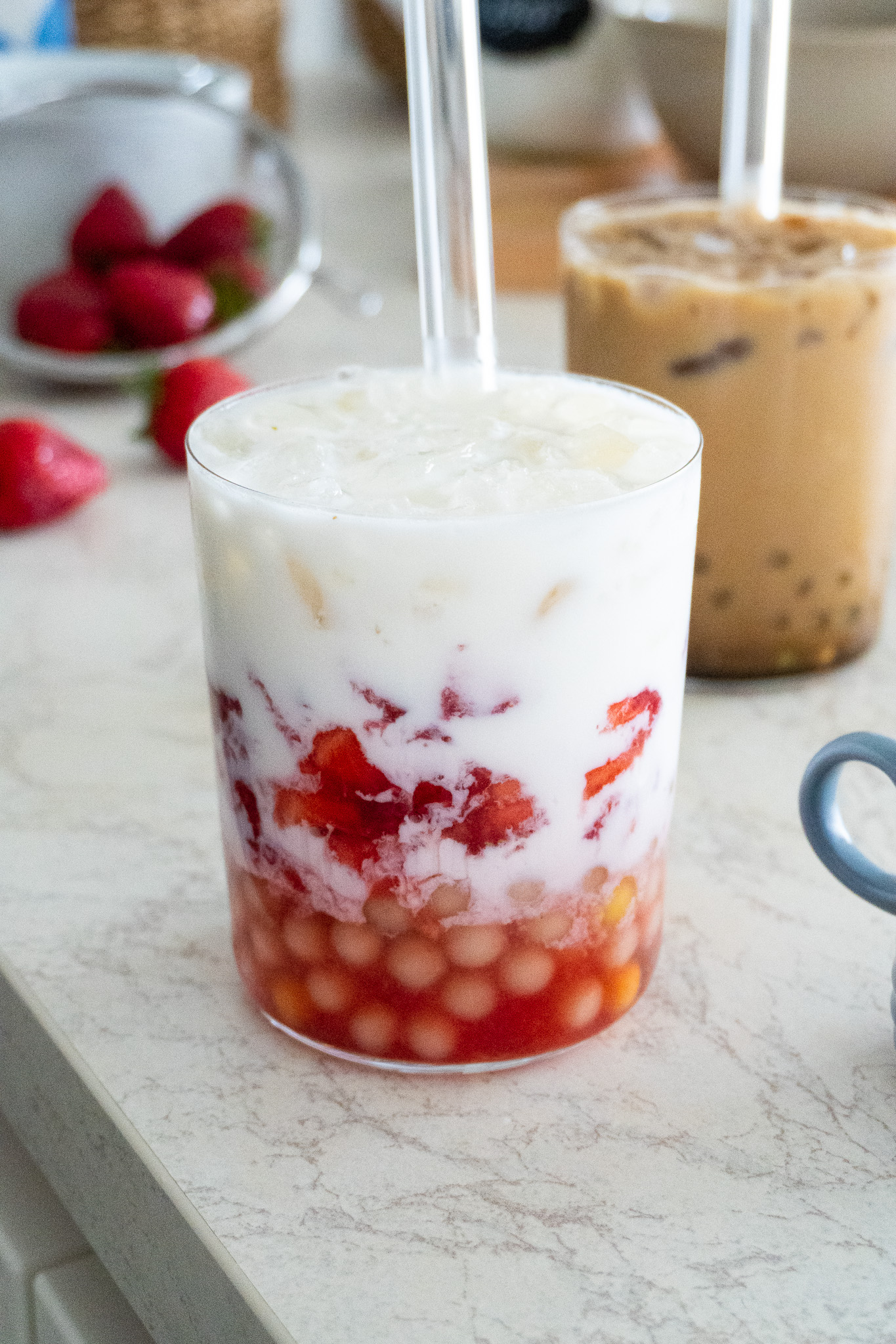 Image of two glasses filled with Easy Homemade Bubble Tea - Korean Strawberry Milk and Brown Sugar Boba with a Shot of Coffee - with tapioca pearls and ice cubes, next to a clear plastic cup with a wide straw.