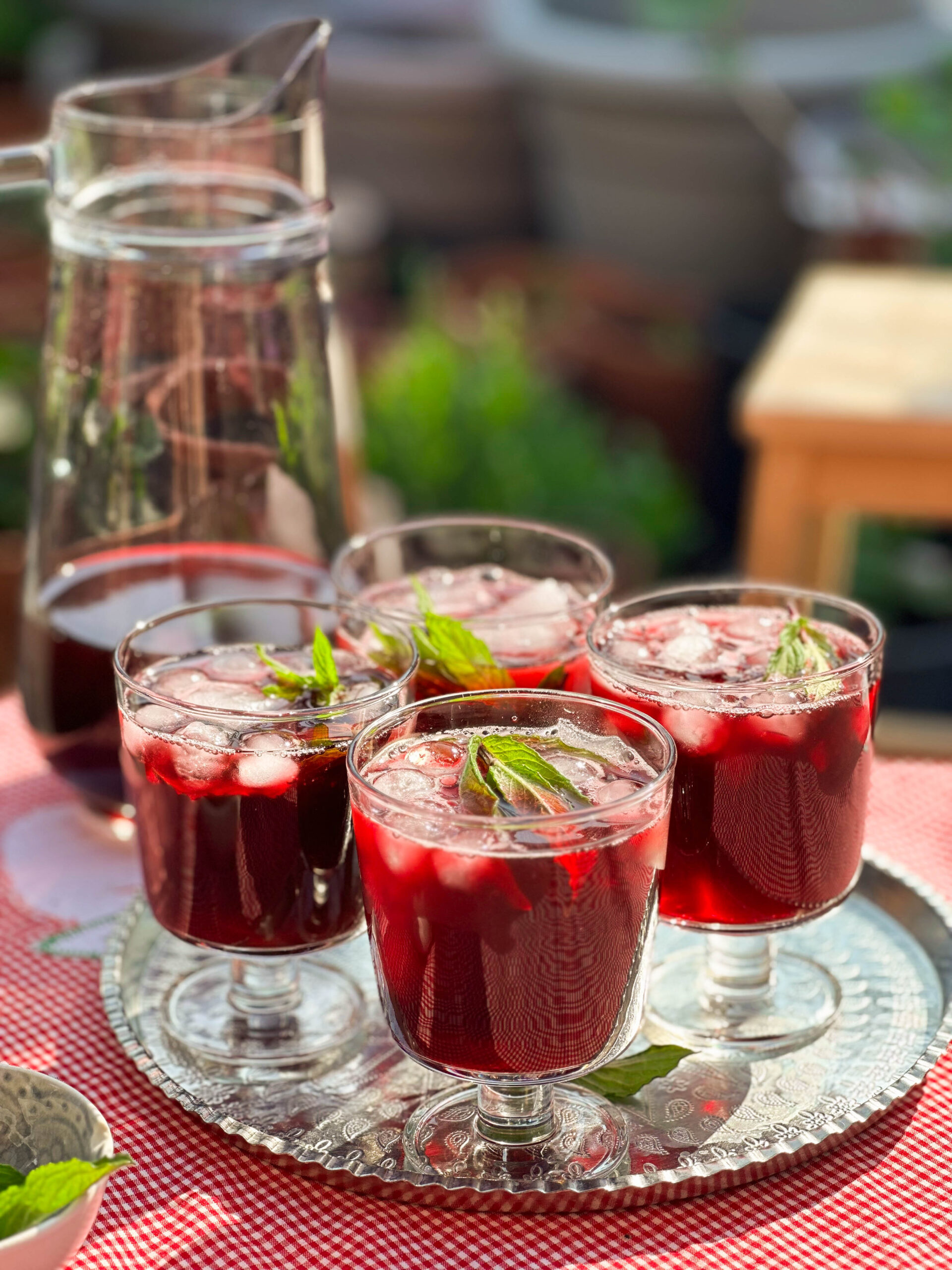 Glass of vibrant red Karkade - Hibiscus Tea on a tray, garnished with fresh mint leaves and ice cubes.