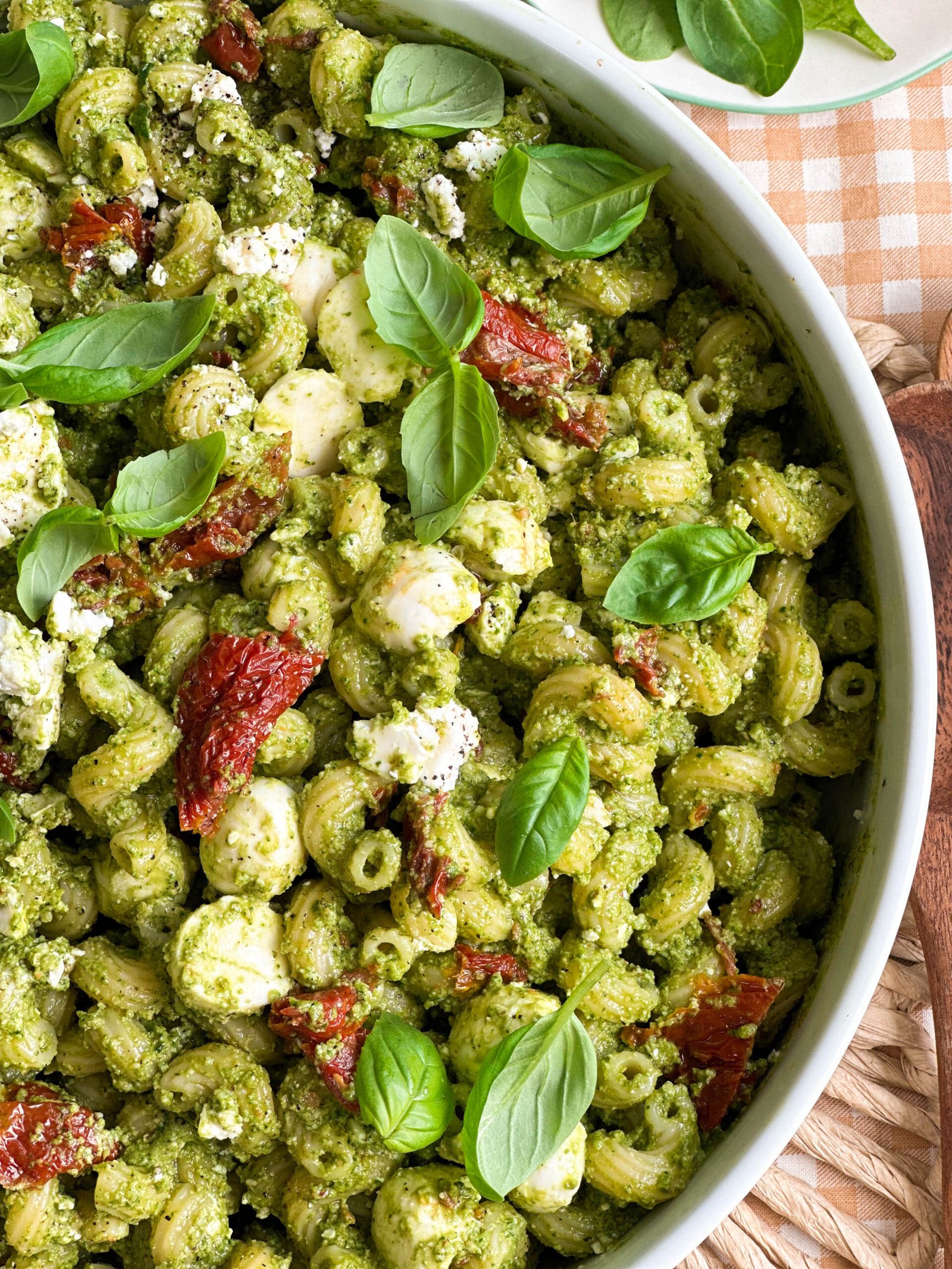 A close-up of a serving of 3 Cheese Spinach Pesto Pasta Salad with sun-dried tomatoes on a white plate, highlighting the colourful ingredients and creamy pesto coating the pasta.