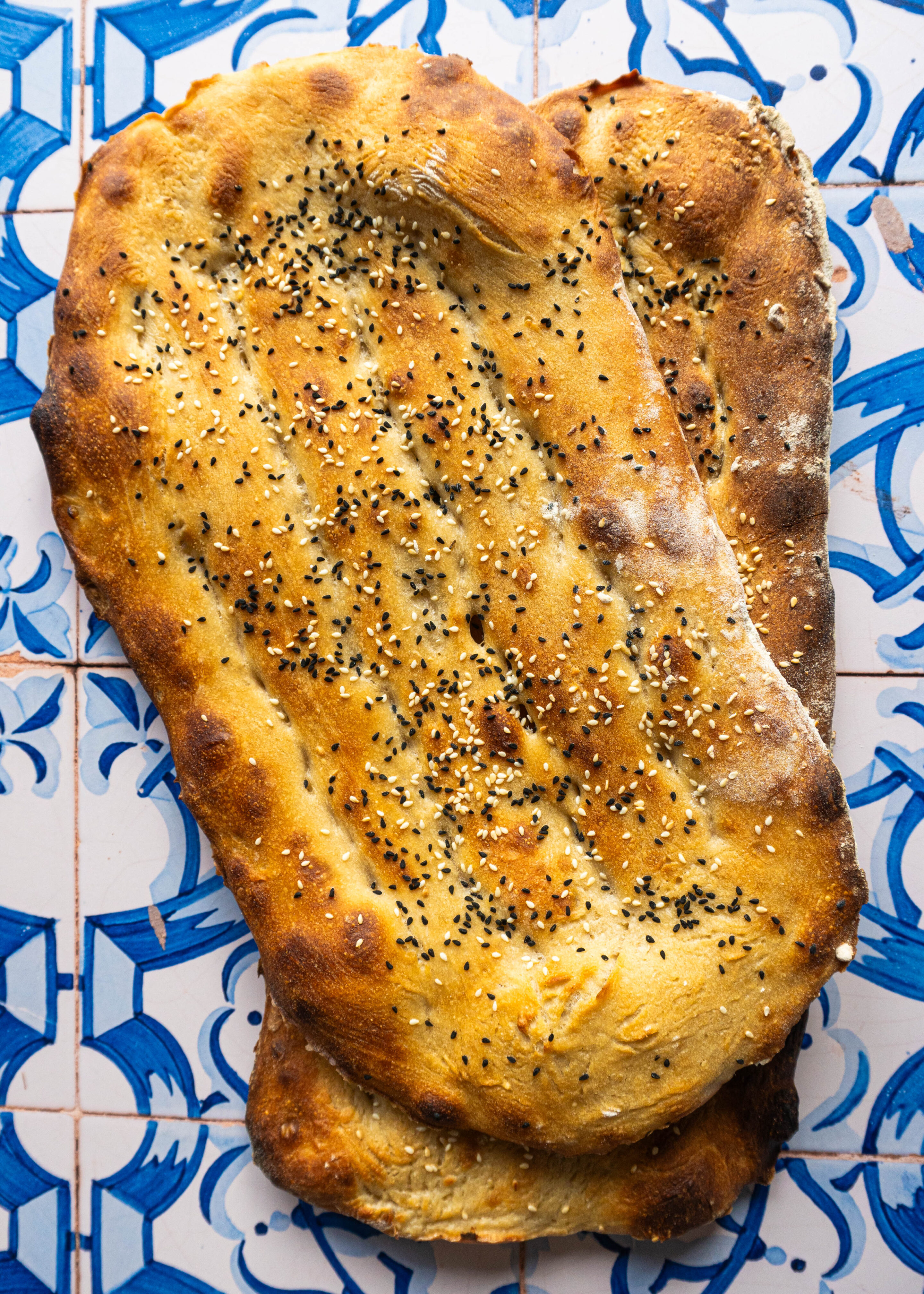 Overhead shot of two freshly baked Persian Barbaris stacked, golden and glistening, sprinkled generously with sesame and nigella seeds, resting on a rustic cloth.