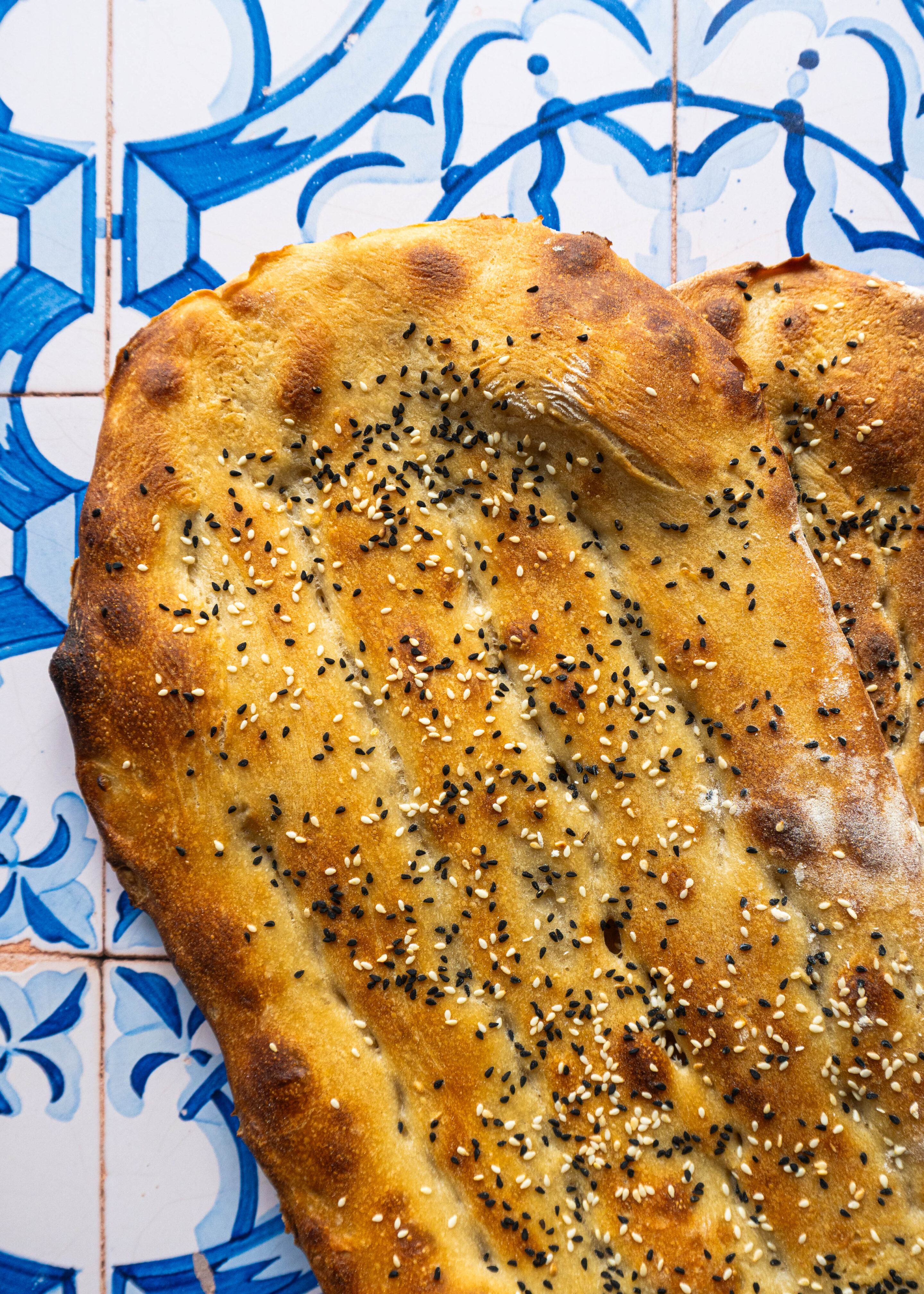 Extreme close-up of a halved Barbari, the crust golden and blistered, with sesame and nigella clinging to its surface, showing off its rich sourdough texture.