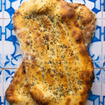 Overhead shot of two freshly baked Persian Barbaris stacked, golden and glistening, sprinkled generously with sesame and nigella seeds, resting on a rustic cloth.