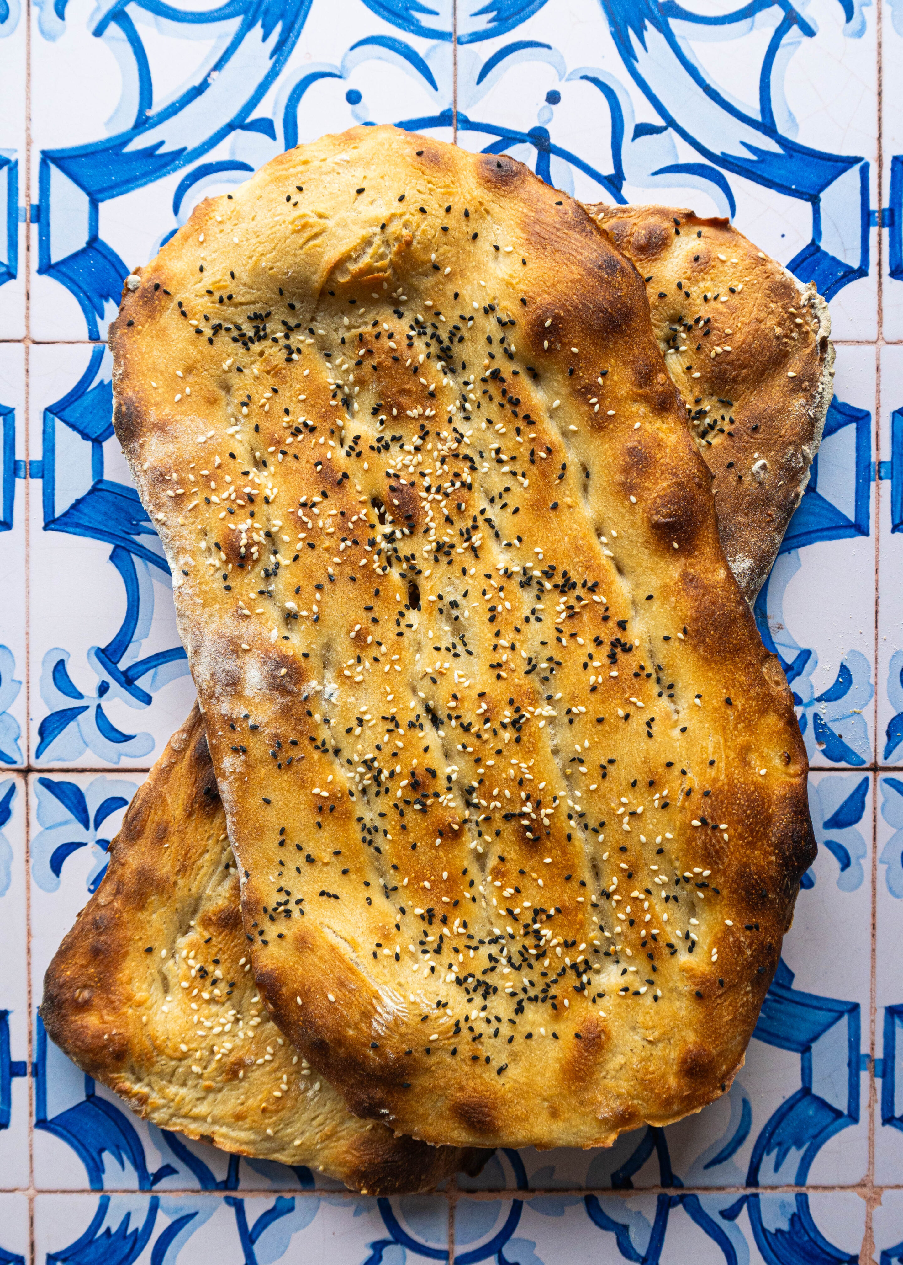 Overhead shot of two freshly baked Persian Barbaris stacked, golden and glistening, sprinkled generously with sesame and nigella seeds, resting on a rustic cloth.