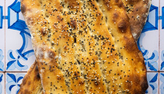 Overhead shot of two freshly baked Persian Barbaris stacked, golden and glistening, sprinkled generously with sesame and nigella seeds, resting on a rustic cloth.