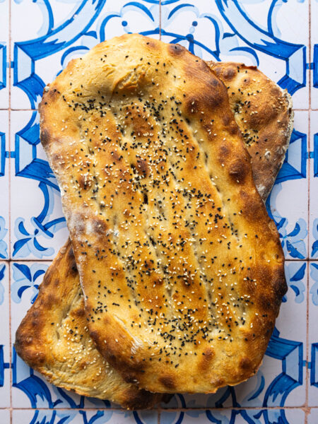 Overhead shot of two freshly baked Persian Barbaris stacked, golden and glistening, sprinkled generously with sesame and nigella seeds, resting on a rustic cloth.