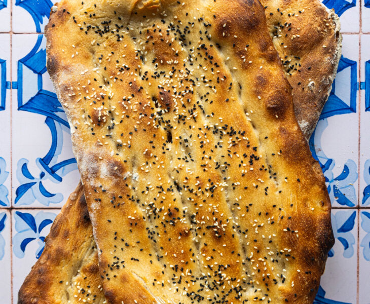 Overhead shot of two freshly baked Persian Barbaris stacked, golden and glistening, sprinkled generously with sesame and nigella seeds, resting on a rustic cloth.