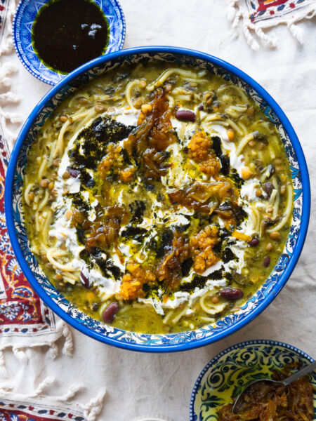 A bowl of Delicious Ashe-Reshteh, a traditional Persian noodle soup with lentils, chickpeas, kidney beans, and fresh herbs, topped with fried onions, mint, and a touch of Kashk.