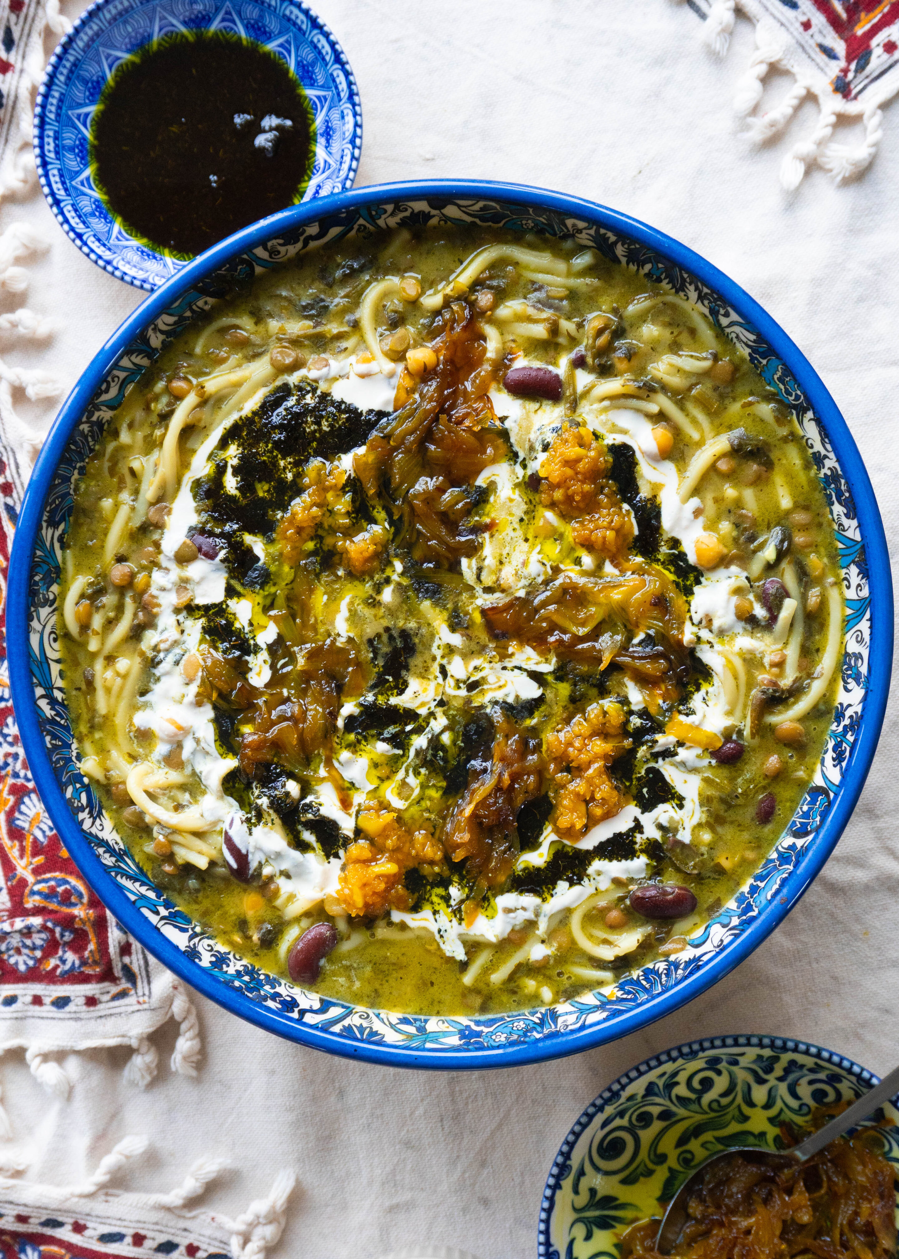 A bowl of Delicious Ashe-Reshteh, a traditional Persian noodle soup with lentils, chickpeas, kidney beans, and fresh herbs, topped with fried onions, mint, and a touch of Kashk.