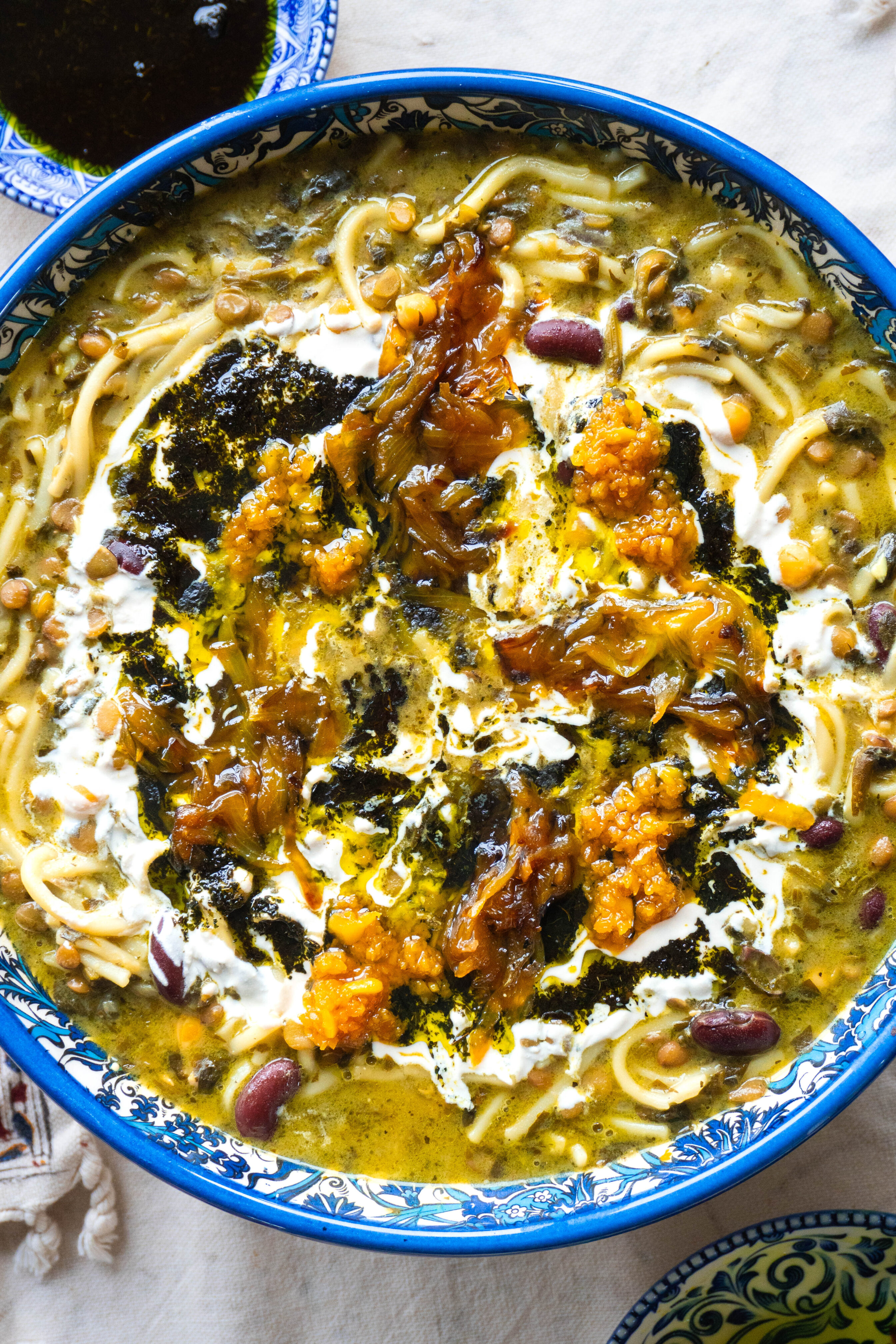 A deliciously aromatic bowl of Ashe-Reshteh, Persian noodle soup with a blend of lentils, herbs, and savory Kashk, garnished with fried onions and fresh mint.