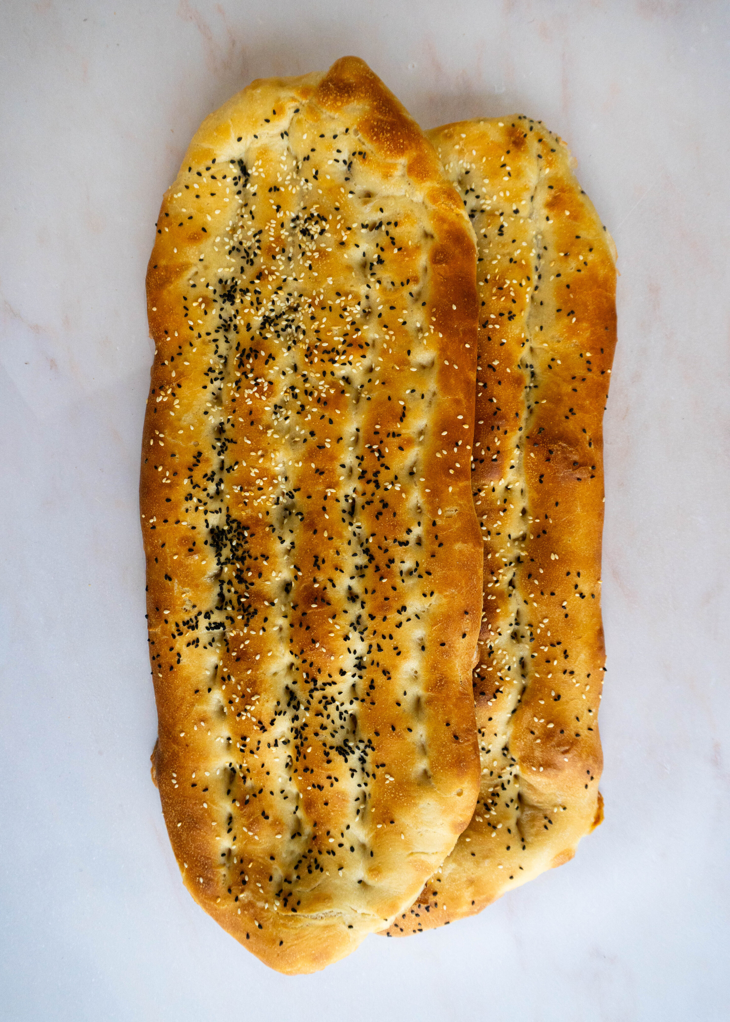 Two oval-shaped Barbari breads, golden brown with deep ridges, sprinkled with sesame and nigella seeds, placed on a white background.