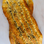 Two freshly baked Barbari breads with a golden crust, topped with sesame and nigella seeds, resting on a light-colored surface.