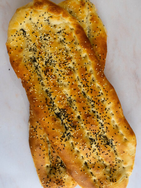 Two freshly baked Barbari breads with a golden crust, topped with sesame and nigella seeds, resting on a light-colored surface.