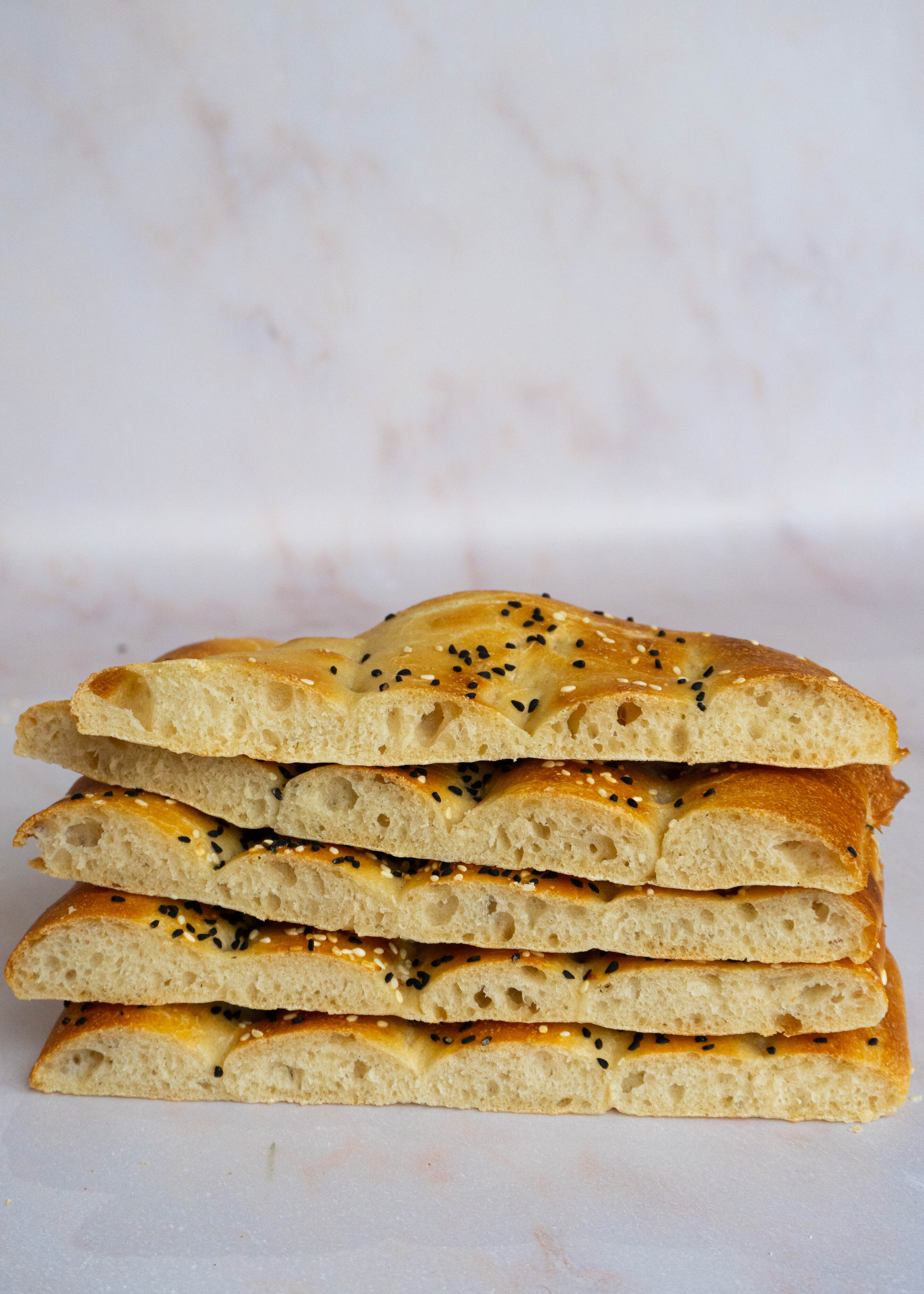 A stack of sliced Barbari bread, showing its airy interior and crisp, golden crust, sprinkled with sesame and nigella seeds.